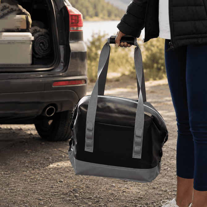 A foldable cooler in black and grey, held by a person near the back of a vehicle. It has carrying straps and a logo.
