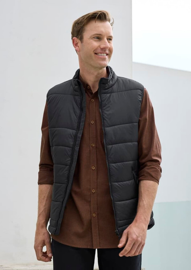 A man wearing a black puffer vest over a brown button-up shirt, smiling in a bright setting.