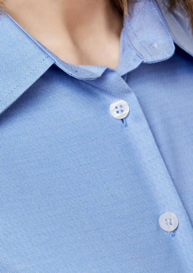 A close-up of a light blue button-up shirt featuring a collar and two buttons. The shirt has a logo.