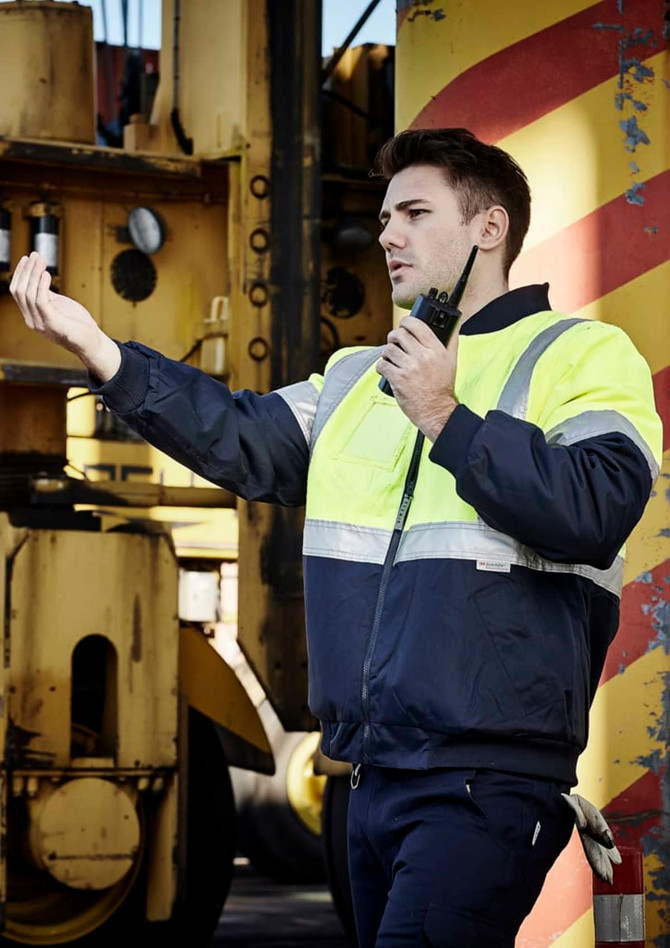 A man in a hi-vis quilted flying jacket, featuring yellow and navy colours, standing with a radio in a work environment.