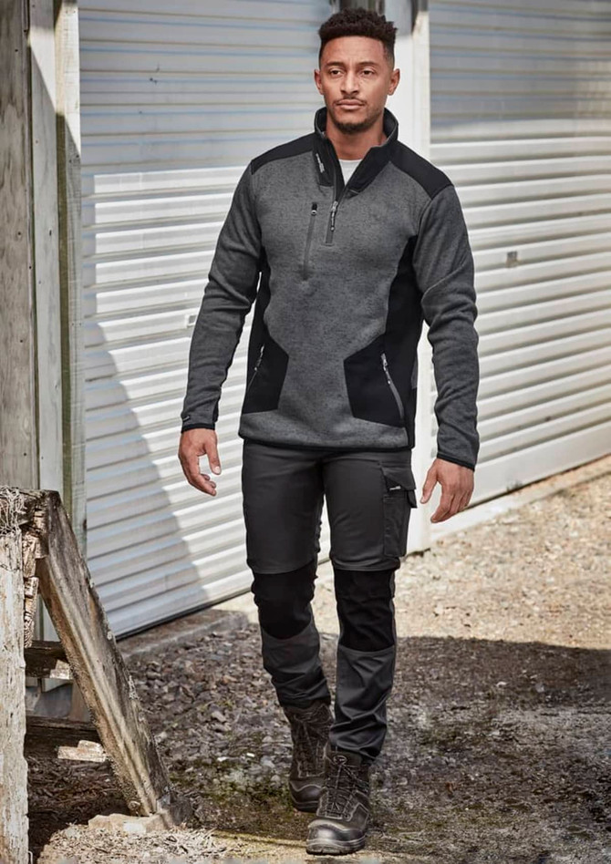 A man in dark grey and black workwear pants and a fitted grey pullover, walking in an outdoor setting.