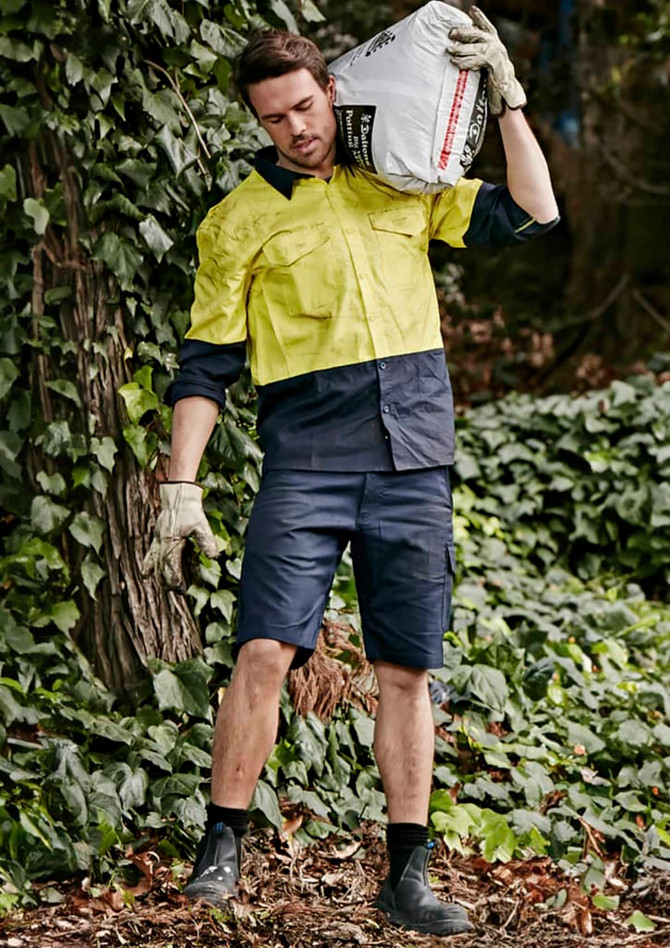 Men's rugged cooling vented shorts in navy, paired with a yellow work shirt, set against a lush green background.