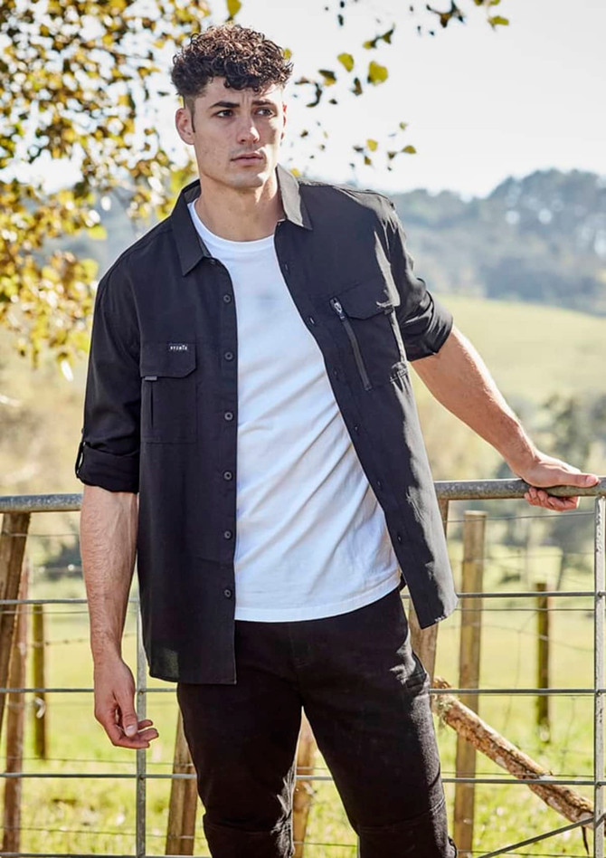 A black long sleeve button-up shirt with pockets, modelled by a man outdoors against a natural backdrop.