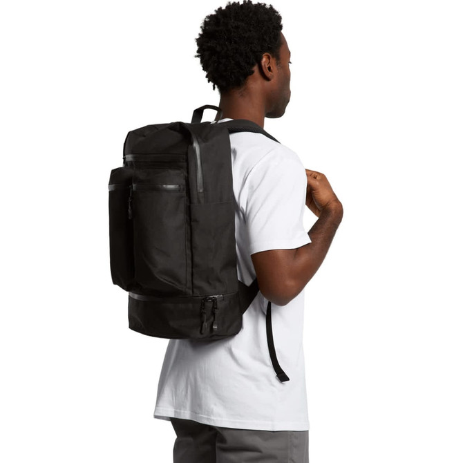 A person wearing a black recycled travel backpack, featuring multiple pockets and a logo on the front.