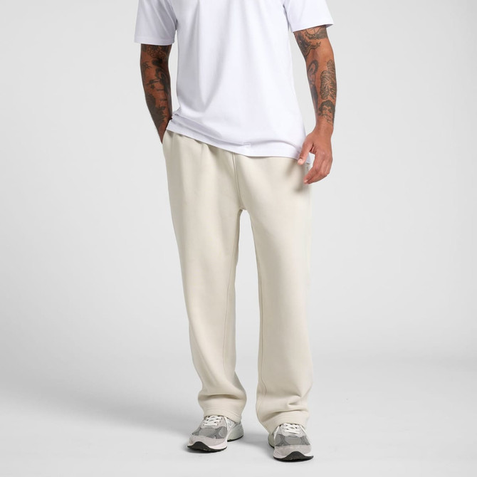 Cuffless track pants in a light beige colour, worn by a person standing with hands in pockets. Features branding on the pants.