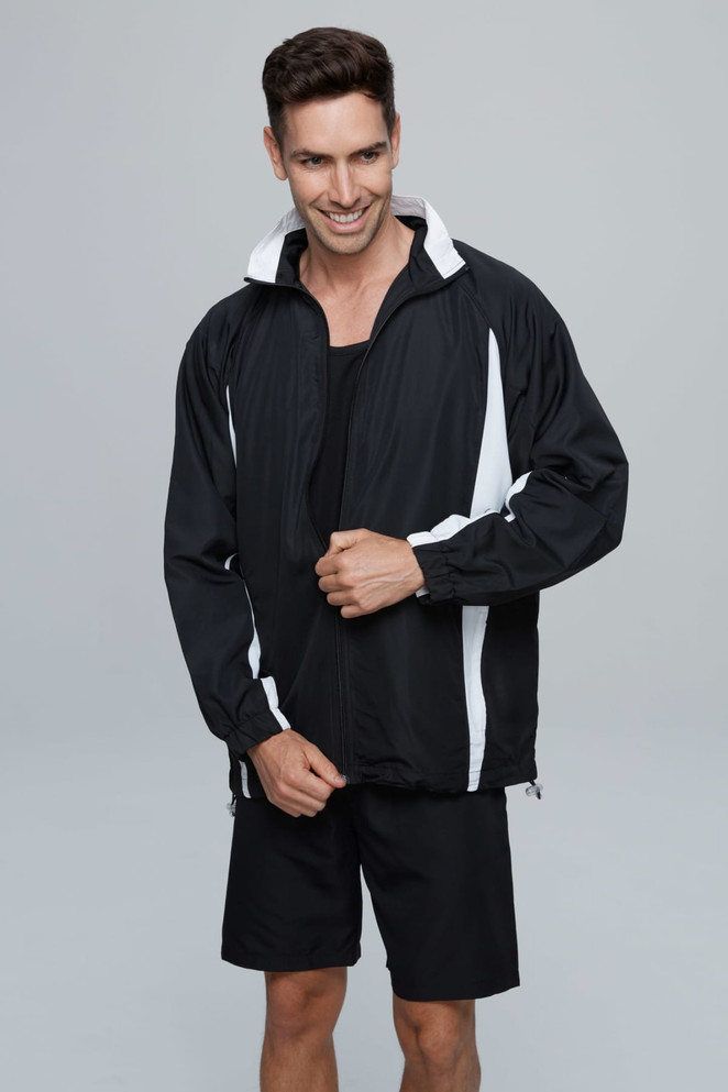 A man in a black track jacket with white stripes, paired with black shorts, standing against a grey background.