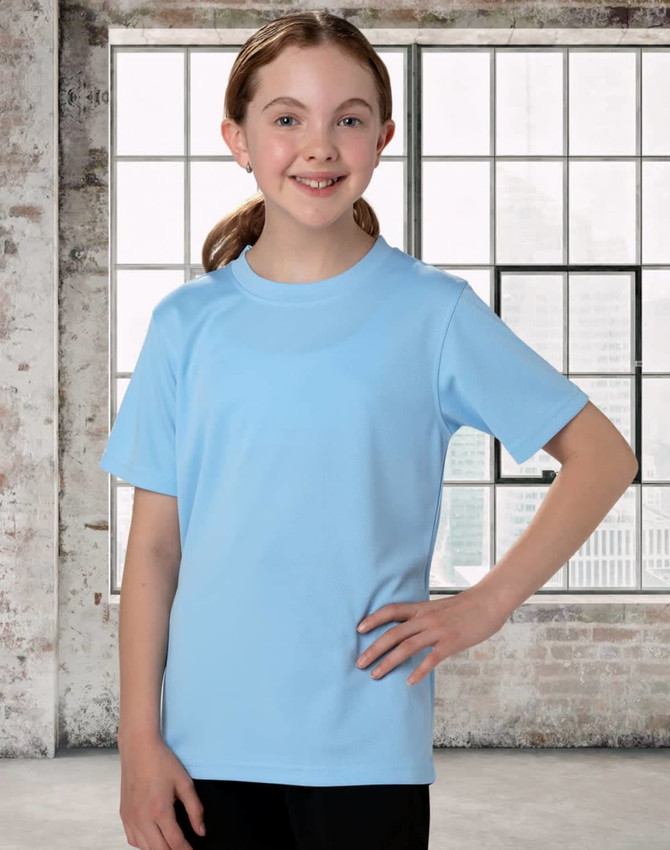 A child in a light blue short sleeve tee, smiling with hands on hips, against a backdrop of large windows.