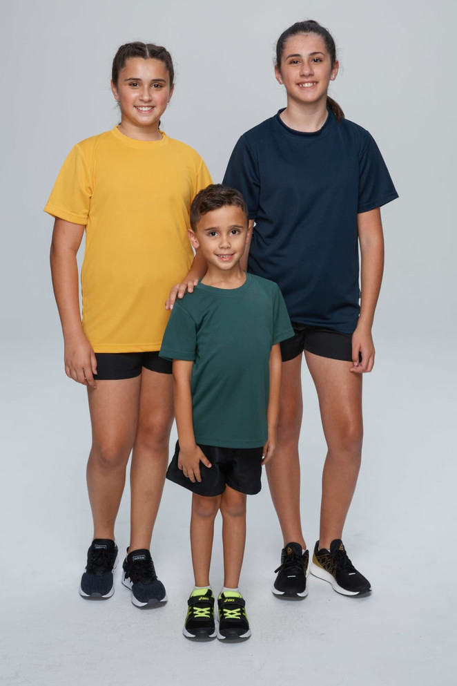 Three children in casual t-shirts: one in yellow, one in dark blue, and one in green. All are wearing shorts and sneakers.