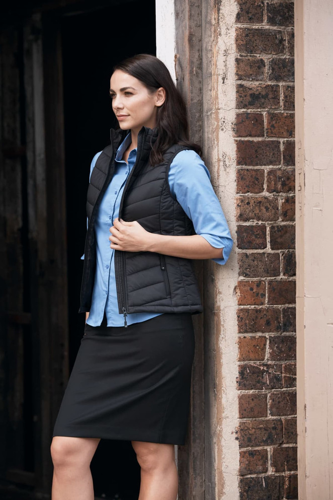 A woman in a black vest stands beside a brick wall, wearing a blue shirt and a black skirt.