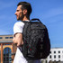 A man wearing a white shirt carries a black Swissdigital Pixel Backpack on his back against a clear blue sky.
