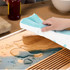 A tea towel in teal and white being used to wipe a wooden surface with water droplets. Features a logo.