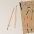 Two wooden grey-lead pencils on a white surface, beside a colourful poster. Each pencil features a logo.