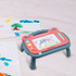 A magnetic drawing board with a stand, featuring grey and pink colours, sits on a light wooden surface beside colourful drawings.