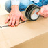 A person sealing a brown cardboard box with black BOPP packing tape, while wearing blue jeans and a light-coloured shirt.