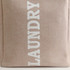 A laundry basket in a beige fabric with white lettering, featuring a sturdy handle and a simple design.