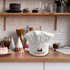 A white chef hat with a logo on a kitchen countertop, surrounded by kitchenware and decorations.