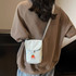 A small canvas shoulder bag in light grey with a flap, carried on a person's shoulder, wearing a brown oversized top.