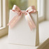 A white gift bag adorned with a soft pink satin ribbon bow, featuring shiny gold lettering.