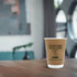 A double wall kraft paper cup in brown, featuring a logo, sits on a wooden table in a modern setting.