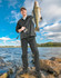 A man stands by the water, holding a large fish and wearing black lightweight nylon waterproof rain pants and a cap.