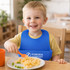 A blue silicone bib with snap closure worn by a smiling child, eating pasta and peas at a dining table.