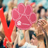 A pink paw foam mitt is raised among a crowd, with hands clapping and cheering in the background.