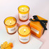 Three amber glass candles with yellow wax, featuring different labels, alongside a small orange box and a decorative ribbon.