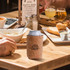 A drink bottle is wrapped in a flat packed cork stubby holder on a wooden table with food and drinks.