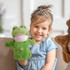 A green frog hand puppet with a smiling face, held by a child wearing a blue dress and a tiara.