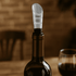 A wine bottle with a clear aerator attached, resting on a wooden surface, with a glass of red wine nearby.