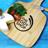 A bamboo cheeseboard with a cheese wedge, cherry tomatoes, leafy greens, and a knife. Features a logo.