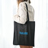 A black shopping tote bag with a V gusset, featuring a logo, held by a person against a light backdrop.