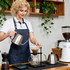 A woman in a navy poly-cotton denim full bib apron with neck strap brews coffee using a kettle and pour-over setup.