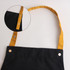 A black poly-cotton canvas apron featuring a yellow colour neck strap and metal snap buttons.