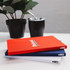 Three A5 notebooks in red, white, and navy stacked on a table with a plant and a drink bottle in the background.