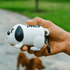 A white dog-shaped waste bag dispenser with black details, held in a hand, featuring a logo.