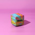 A colourful, rainbow-patterned cube featuring textured bumps, set against a pink background, with a logo.