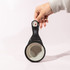 A hand holds a black collapsible silicone pet food scoop with a grey interior and a logo on the handle.