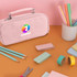 A pink pencil case with compartments and a handle, surrounded by stationery items like coloured pencils and tape.