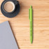 A green Wheat Straw ballpoint pen placed on a wooden surface next to a black coffee cup and grey paper.