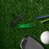 A golf divot tool in black and green, alongside golf tees, a golf ball, and a club, on a grass-like surface.