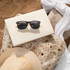 Bamboo sunglasses with black frames and dark lenses resting on a light-coloured box, surrounded by rocks and a straw hat.