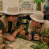 Two children wearing beige PVC safari hats sit together, examining a map amidst a decorated jungle-themed classroom.