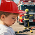 A child wearing a shiny red PVC fire chief hat stands in front of a fire truck, with a firefighter in the background.