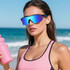 A woman wearing blue mirrored sports cycling sunglasses and a pink tank top, holding a pink drink bottle by the beach.