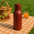 A drink bottle in a matte red finish, with a sleek design and a logo, placed on a picnic blanket.