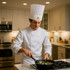 A chef in a white disposable non-woven chef hat cooks with a pan in a modern kitchen. The hat features a logo.