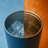 A stainless steel coffee mug, with one side showcasing ice and the other side filled with steaming liquid. It has a logo.
