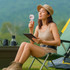A woman sits in a camping chair, holding a pink transparent fan with LED light, surrounded by greenery.