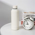 A drink bottle in a sleek off-white colour beside a vintage alarm clock and a stack of books.